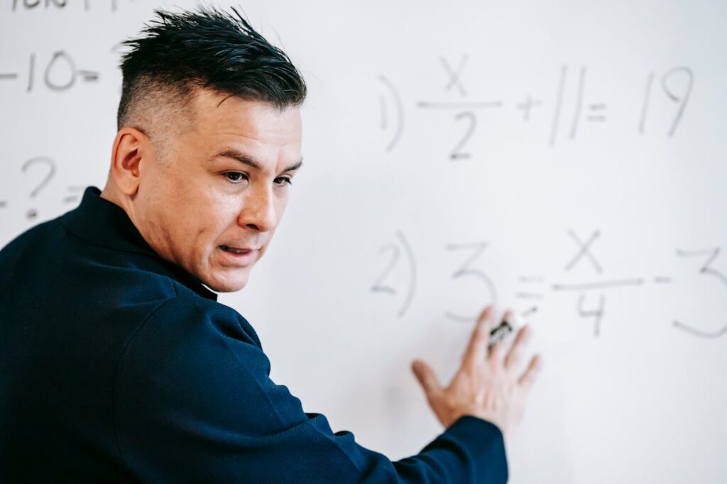 A mathematics instructor explaining algebraic equations on a whiteboard in a classroom setting.